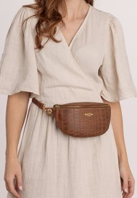 Borsa a cintura in ecopelle di coccodrillo marrone con forma rotonda, hardware dorato e chiusura a zip. Indossata in vita sopra un vestito beige chiaro.