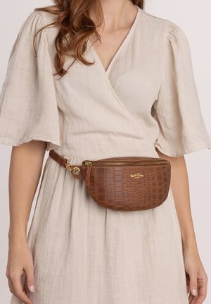 Borsa a cintura in ecopelle di coccodrillo marrone con forma rotonda, hardware dorato e chiusura a zip. Indossata in vita sopra un vestito beige chiaro.