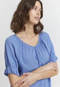 Blusa blu a maniche corte con scollo a V, texture arricciata e polsini volant. Tessuto leggero con una caduta morbida, senza motivi visibili.