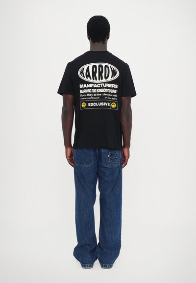 Tricou din bumbac negru, cu text alb mare pe spate, însoțit de elemente de design grafic și logo-uri. Asortat cu blugi din denim albastru deschis.