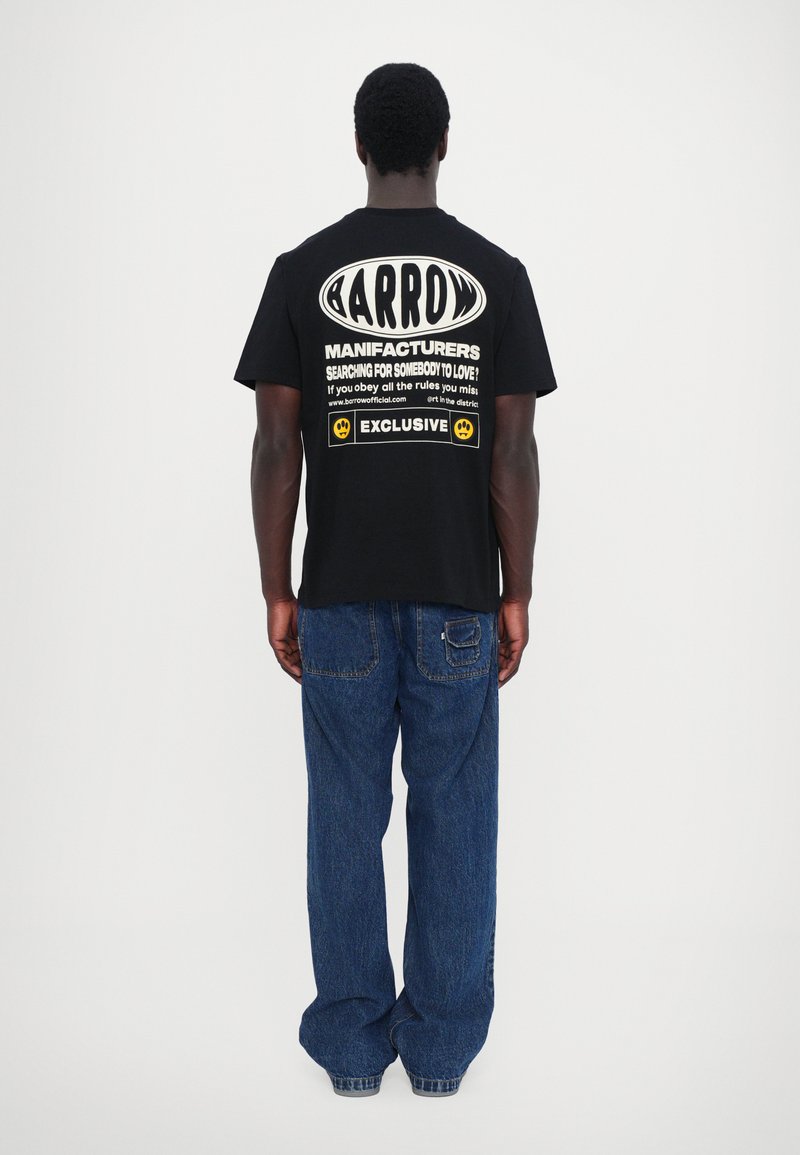 Camiseta de algodón negra con gran texto blanco en la parte posterior, con elementos de diseño gráfico y logotipo. Combinada con jeans de mezclilla azul claro.