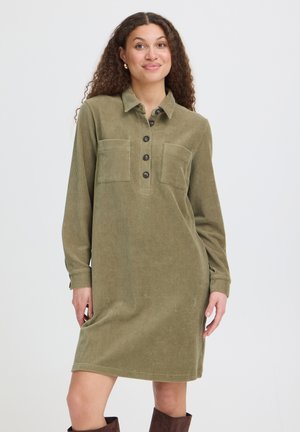 Vrouw met krullend haar die een groene corduroy jurk met knopen en bruine laarzen draagt, staand met één hand in de zak tegen een effen achtergrond.
