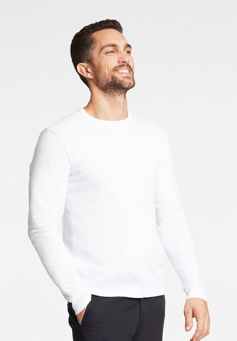 Weißes Langarm-T-Shirt mit Rundhalsausschnitt und figurbetontem Schnitt, kombiniert mit dunkler Hose, vor einem schlichten Hintergrund präsentiert.