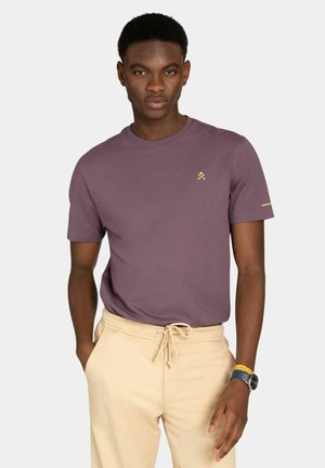 Jeune homme portant un t-shirt mauve avec un petit logo sur la poitrine et un pantalon beige à cordon, debout avec une main dans la poche.