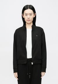 Melna džemperis ar rāvējslēdzēju, stāvošu apkakli, ribveida aprocēm un nelielu zīmola emblēmu uz krūtīm. Valkāts virs baltas krekla.