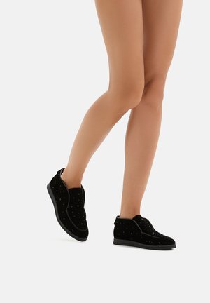 Helle Beine in schwarzen Slippers mit kleinen dekorativen Nieten, ein Fuß leicht angehoben, vor einem weißen Hintergrund.