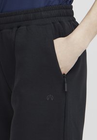 Čierne tepláky s elastickým pásom, bočnou vrecko a detailom na šnúrku. Obsahujú malé embosované logo blízko vrecka. Hladká textúra.