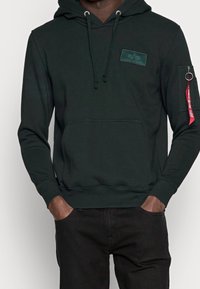 Felpa verde scuro realizzata in morbido tessuto, con tasca a marsupio, cappuccio con coulisse e tasca a zip sulla manica con dettaglio di etichetta rossa.