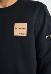 Sudadera azul marino de algodón, con una etiqueta grande color canela que dice "Columbia Sportswear Company" y un pequeño logotipo en la manga.