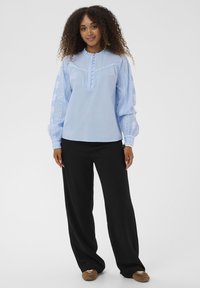 Blouse bleu clair en tissu doux, avec un devant plissé, des boutons et des accents brodés sur les manches bouffantes. Associée à un pantalon noir.