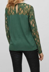 Blusa verde con maniche lunghe in pizzo, caratterizzata da un motivo floreale trasparente. Tessuto del corpo liscio, dettaglio di apertura a chiave sul retro, abbinata a leggings neri testurizzati.