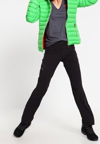 Zielona kurtka puchowa, szary t-shirt z dekoltem w serek oraz czarne spodnie outdoorowe. Kurtka ma pikowany wzór i zamki błyskawiczne; spodnie są dopasowane i lekkie.