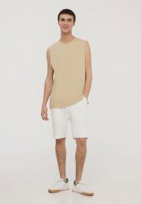 Top senza maniche beige abbinato a pantaloni corti bianchi arrotolati. La modella indossa scarpe da ginnastica bianche con accenti verdi. Sfondo neutro.