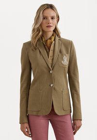 Blazer marron sur mesure avec deux poches avant, boutons en argent et détail brodé sur la poche, porté sur une chemise beige et un pantalon rose.