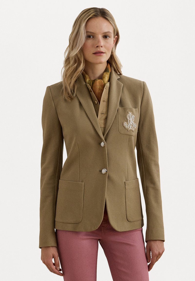 Blazer marron sur mesure avec deux poches avant, boutons en argent et détail brodé sur la poche, porté sur une chemise beige et un pantalon rose.