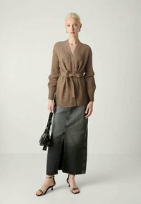 Καφέ πλεκτό cardigan με ζωνάκι, συνδυασμένο με φούστα τζιν μέχρι το γόνατο και μαύρα σανδάλια με λουράκια; μοντέλο κρατάει μια μικρή μαύρη τσάντα.