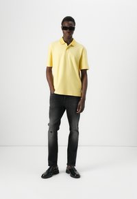 Gul polo-skjorte med krage, korte ermer, kombinert med falmede svarte jeans og svarte sko. Modellen står mot en enkel bakgrunn.