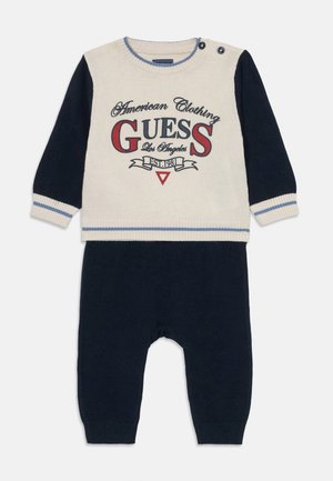 Knitted two-piece baby outfit in cream and navy. Features printed text, ribbed cuffs, and button accents on the shoulder. Soft texture.