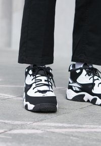 Osoba nosící černo-bílé robustní tenisky s černými tkaničkami, stojící na kamenné dlažbě, viditelná od poloviny lýtek dolů.