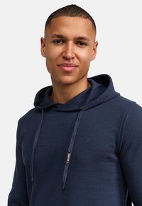 Dunkelblaues, geripptes Kapuzenpullover aus strukturiertem Stoff. Verfügt über eine Kapuze mit Kordelzug und ein kleines Logo-Emblem auf der Brust.