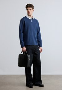 Μπλε σκούρο φούτερ με γιακά και λευκό πουκάμισο από μέσα, συνδυασμένο με σκούρο τζιν παντελόνι φαρδύ και μαύρη δερμάτινη τσάντα.