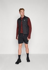 Sort quilted vest med høj krave, båret over en mørkerød langærmet skjorte. Sorte shorts og sko fuldender den atletiske påklædning.