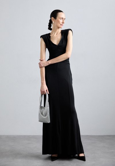 Largo vestido negro con mangas tipo bolsillo y cuerpo fruncido; combinado con un bolso de mano joya plateado y tacones negros de punta. Diseño simple y elegante.