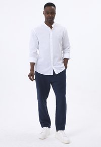 Camisa blanca de manga larga con cuello de banda, combinada con pantalones azul marino y zapatillas blancas, con un diseño minimalista y tejido texturizado.