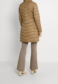 Brun puffertjacka med ett chevronmönster, knälång, hög krage och långa ärmar. Bärs med beige byxor och klackade ankelstövlar.