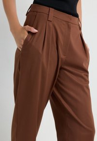 Persoon in high-waisted bruine geplooide broek met de handen in de zakken, gecombineerd met een ingestopt zwart topje tegen een effen achtergrond.