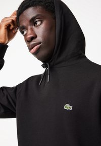 Jeune homme portant un sweat à capuche noir avec un logo de crocodile vert, touchant sa tête et regardant sur le côté sur un fond blanc uni.