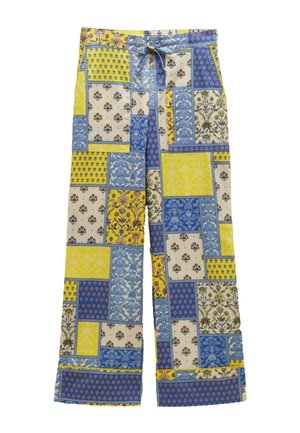 Jeune femme en T-shirt court blanc, pantalon large patchwork bleu et jaune, collier de perles bleues et tongs jaunes, debout contre un mur beige.