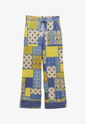 Jeune femme en T-shirt court blanc, pantalon large patchwork bleu et jaune, collier de perles bleues et tongs jaunes, debout contre un mur beige.