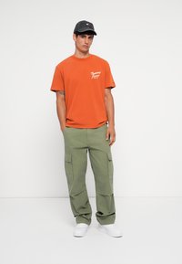 Orange cotton t-shirt with white logo, paired with green cargo pants featuring side pockets, and white sneakers. Black cap completes the look.