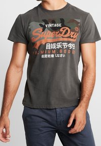 Šedé bavlněné tričko s maskáčovým vzorem na horní části přední strany. Obsahuje oranžové logo "SuperDry" a japonský text. Krátké rukávy.