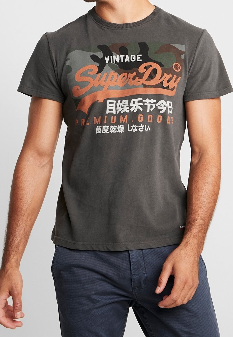 Šedé bavlněné tričko s maskáčovým vzorem na horní části přední strany. Obsahuje oranžové logo "SuperDry" a japonský text. Krátké rukávy.