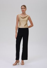 Top de crema satinado con escote tipo cowl, combinado con pantalones negros con acentos laterales. La modelo lleva tacones de tiras en color beige.