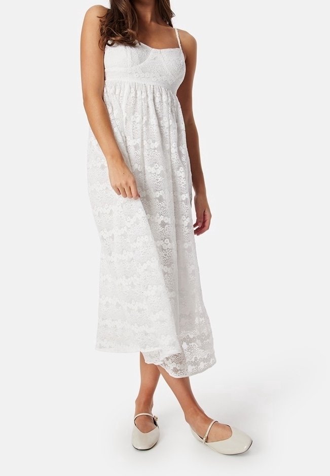 Femme portant une robe midi en dentelle blanche avec des bretelles spaghetti et des ballerines Mary Jane beiges, debout devant un fond uni.