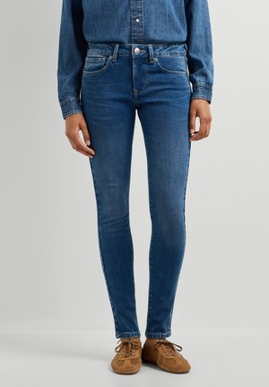 Persoană îmbrăcată cu blugi albaștri mulați, cămașă de denim albastră asortată și pantofi casual maro cu șireturi, stând în fața unui fundal simplu.