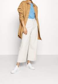 Camisa beige de botones, debajo una camisa a rayas azul y blanca, pantalones anchos blancos y zapatillas blancas, con un corte relajado y tejido suave.