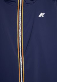 Giubbino antivento blu navy con una cerniera bianca che presenta strisce rosse, gialle e nere. Include un logo bianco sul lato sinistro. Texture liscia.