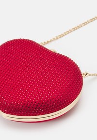 Czerwona torebka w kształcie serca udekorowana małymi czerwonymi cyrkoniami i złotym paskiem na białym tle.