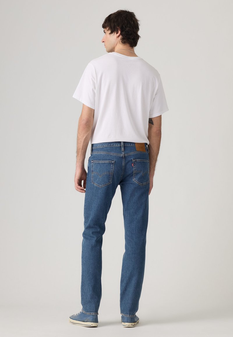 Hombre con camiseta blanca de manga corta, vaqueros azules y zapatillas azules, de pie de espaldas sobre un fondo liso gris claro.
