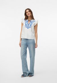 Witte korte mouwen top met blauwe bloemenborduursels en een V-hals, gecombineerd met lichtblauwe wijde jeans en donkerblauwe sandalen.