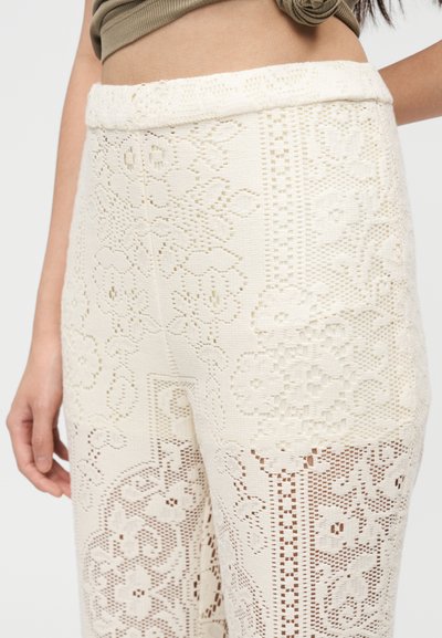 Γυναίκα με ψηλόμεσα, κρεμ δαντελένια παντελόνια με λουλουδένια μοτίβα και κοντό ελιάς τοπ μπροστά σε απλό φόντο.