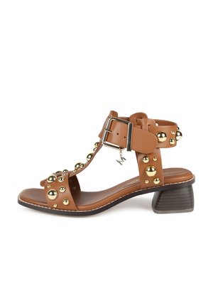 Sandale en cuir marron avec des clous dorés, design en T, et un petit talon carré. Présente une boucle en argent et des détails de couture minimaux.