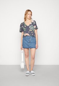 T-shirt floral em cinza escuro com decote em V e mangas curtas, combinado com uma saia de ganga azul clara. Os acessórios incluem uma mala branca e ténis cinzentos.