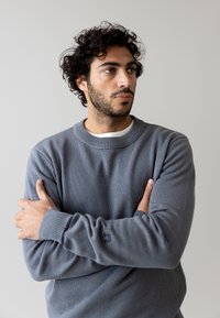 Suéter gris de punto con cuello redondo, puños acanalados y un sutil logo en la manga. La tela tiene un acabado texturizado.