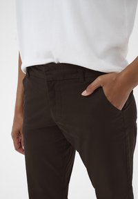 Sötétbarna pamut nadrág sima textúrával, slim fit szabással, elülső zsebekkel és közepes derékvonallal. Egy fehér felsővel párosítva.