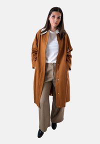 Bruine lange jas met drukknopen, witte kraag en voering, gedragen over een wit shirt en beige gestructureerde wijde broek. Zwarte enkelhoge laarzen.